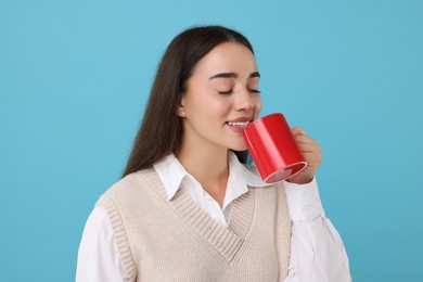 Happy young woman drinking beverage from red ceramic mug on light blue background