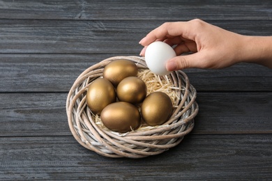 Photo of Woman taking egg from nest with gold ones on wooden table