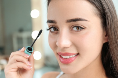 Photo of Attractive young woman applying mascara on her eyelashes against blurred background