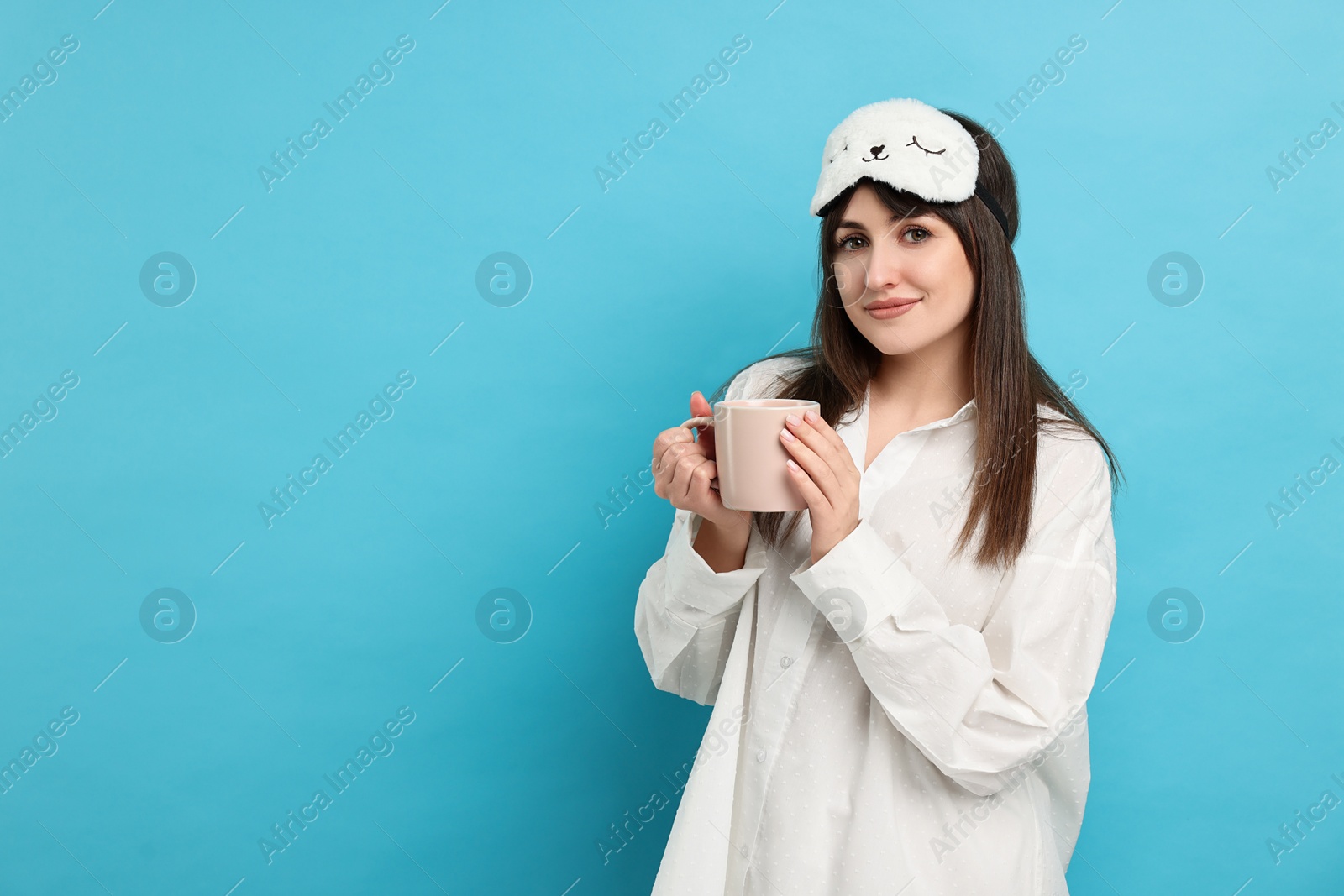 Photo of Woman in pyjama and sleep mask holding cup of drink on light blue background, space for text