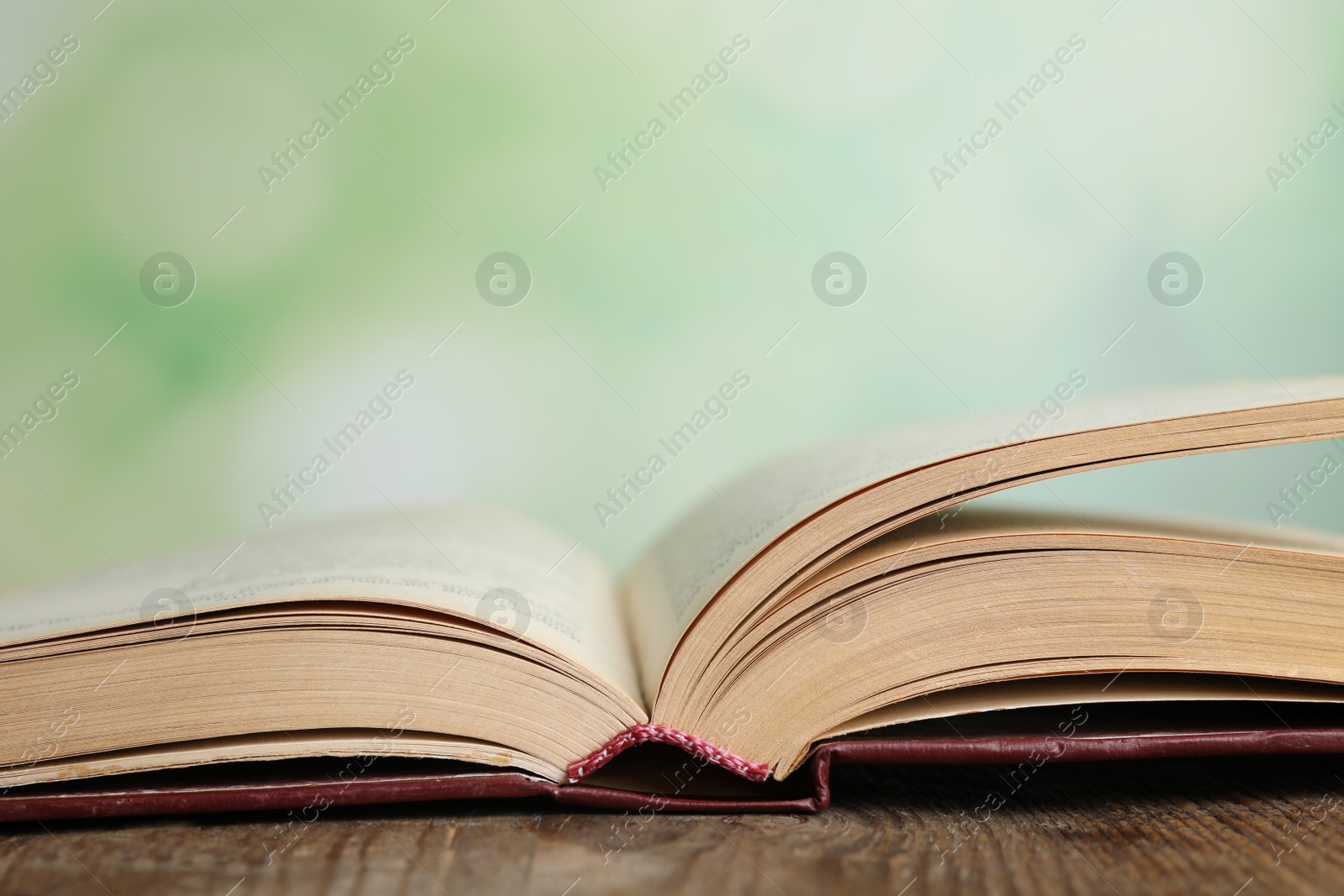 Photo of Open book on wooden table against blurred green background, closeup. Space for text