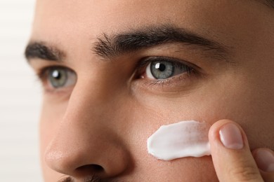 Man with dry skin applying cream onto his face on light background, closeup