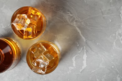 Photo of Whiskey with ice cubes in glasses and bottle on light grey textured table, flat lay. Space for text