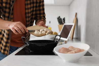 Photo of Man putting cut mushrooms into frying pan while watching online cooking course via laptop in kitchen, closeup