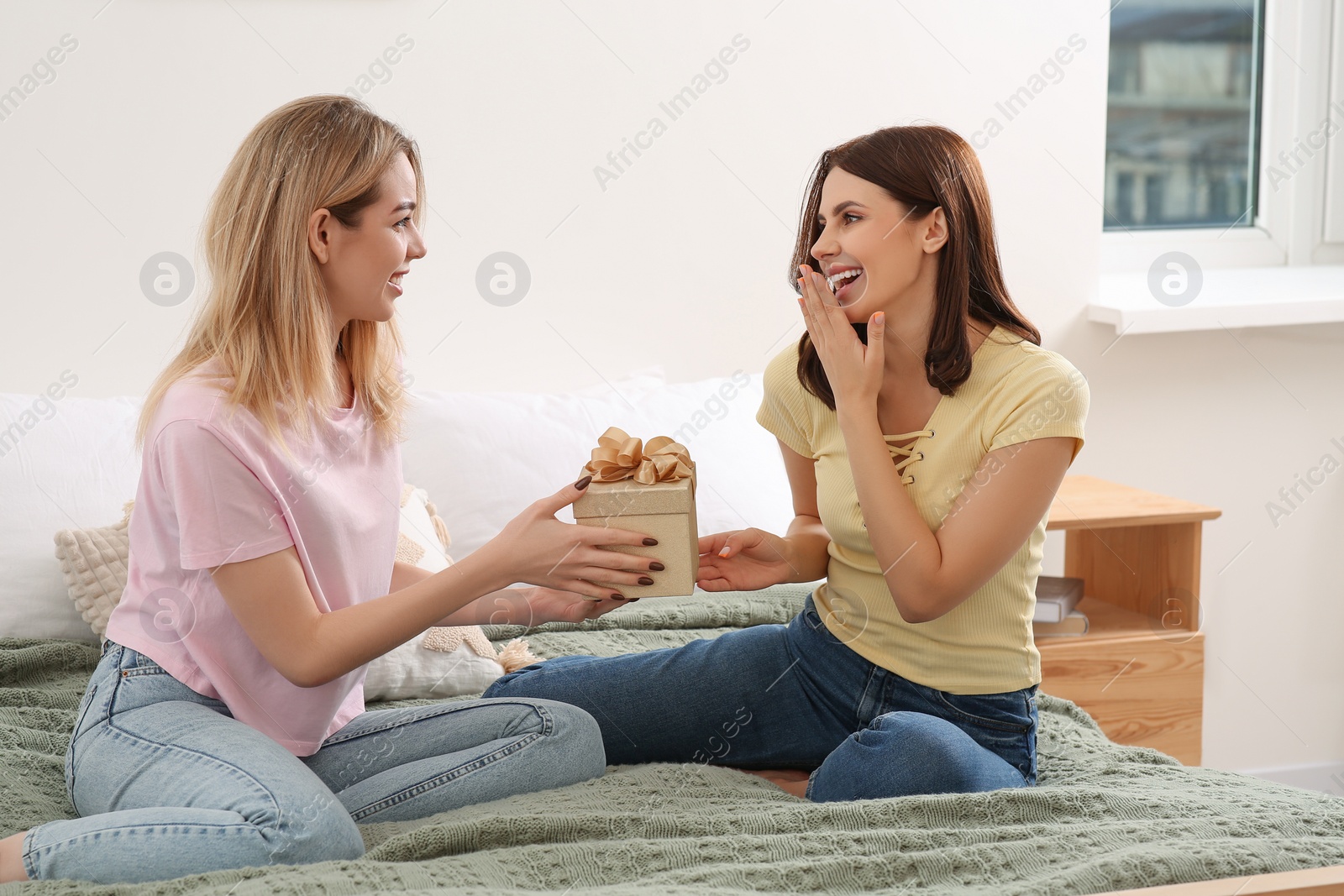 Photo of Smiling young woman presenting gift to her friend on bed at home