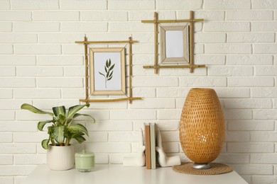 Potted houseplant, decor elements and books on white table near brick wall with stylish bamboo frames