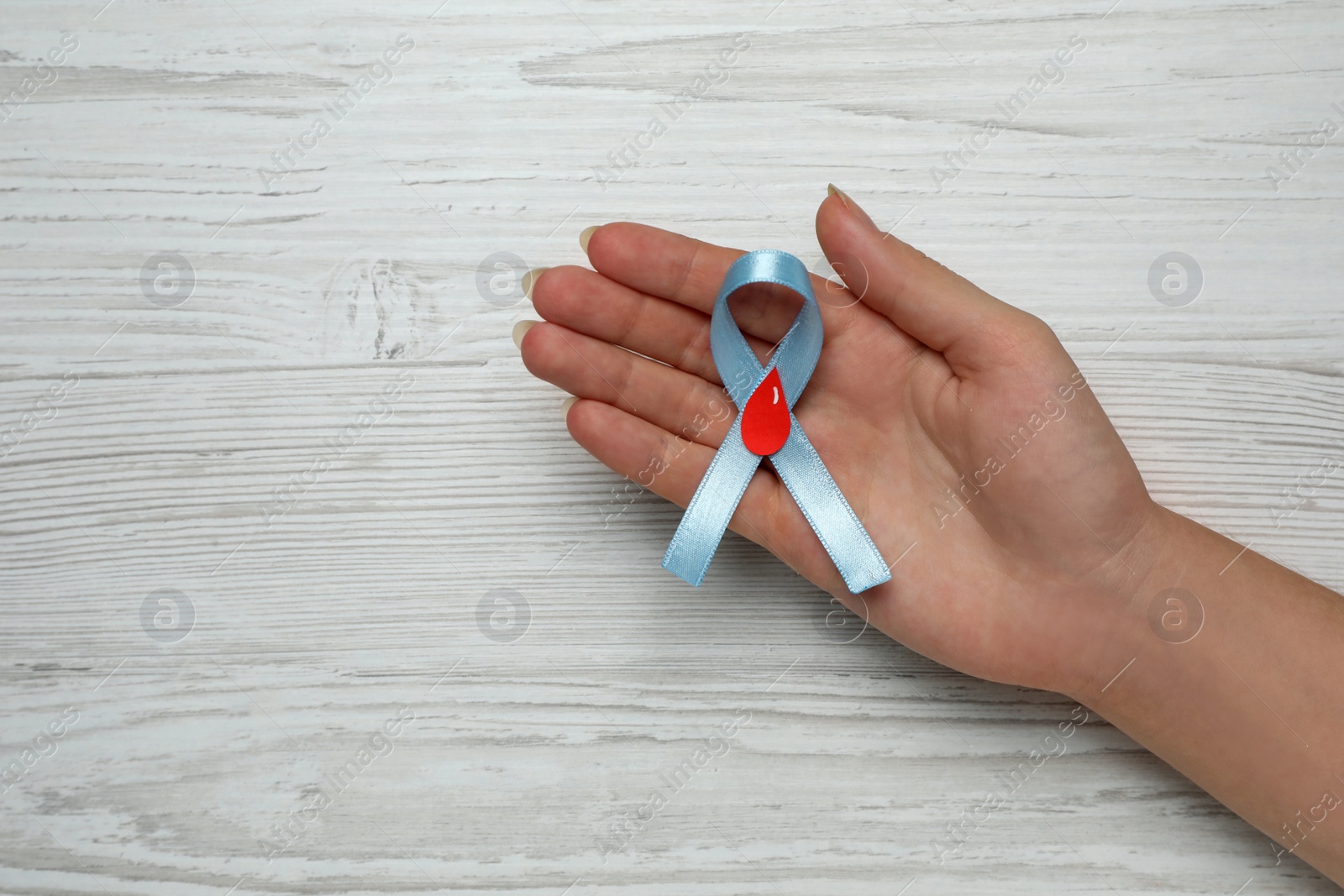 Photo of Woman holding light blue ribbon with paper blood drop at wooden table, top view and space for text. Diabetes awareness