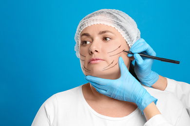 Doctor drawing marks on woman's face for cosmetic surgery operation against blue background. Double chin problem