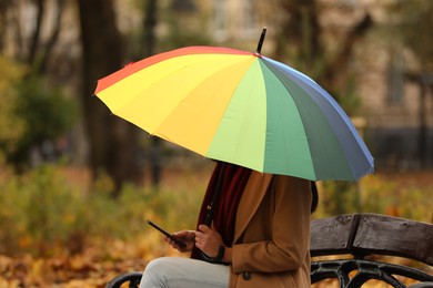 Photo of Woman with rainbow umbrella and smartphone sitting on bench in autumn park