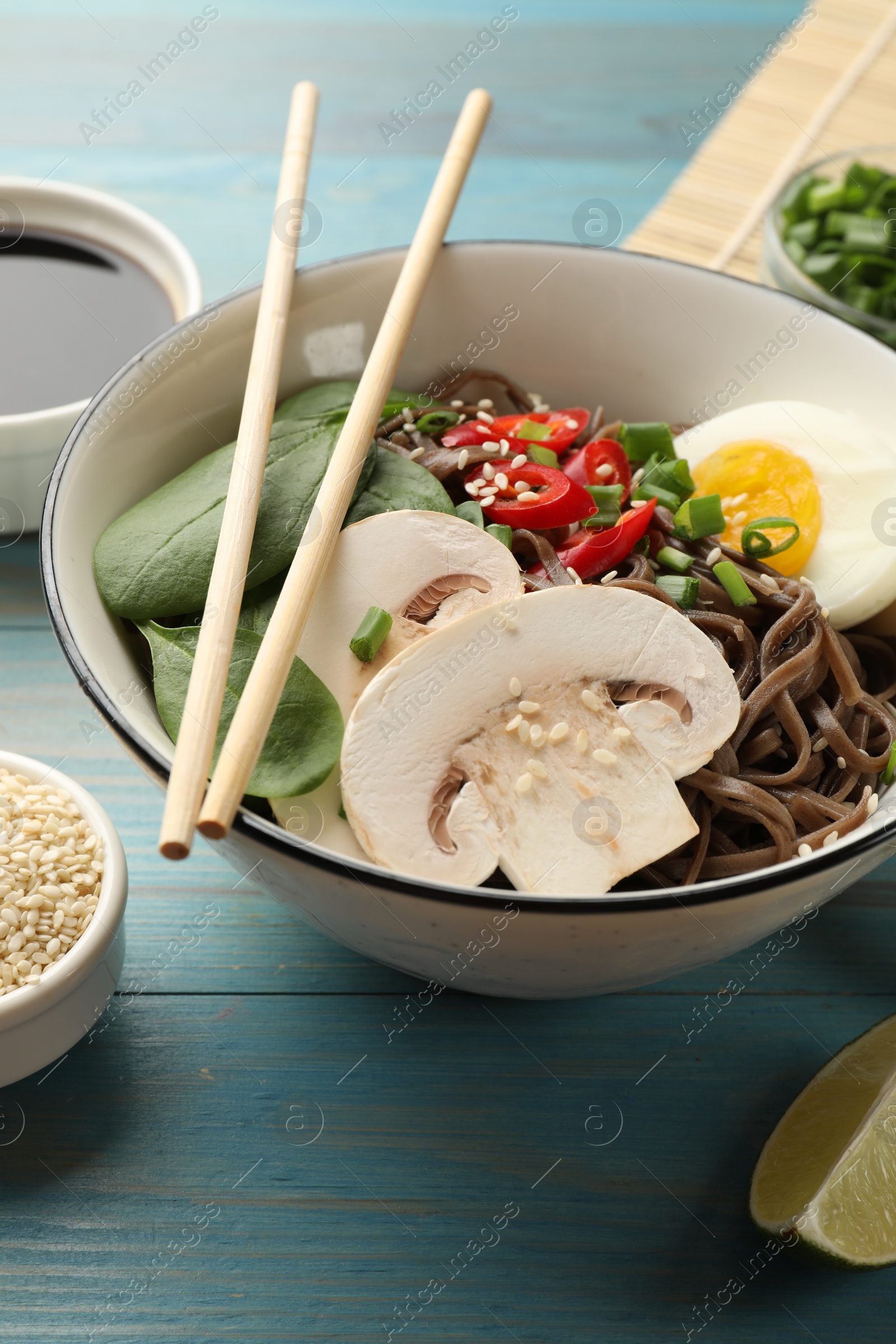 Photo of Tasty buckwheat noodles (soba) with chili pepper, egg, mushrooms and chopsticks on light blue wooden table, closeup