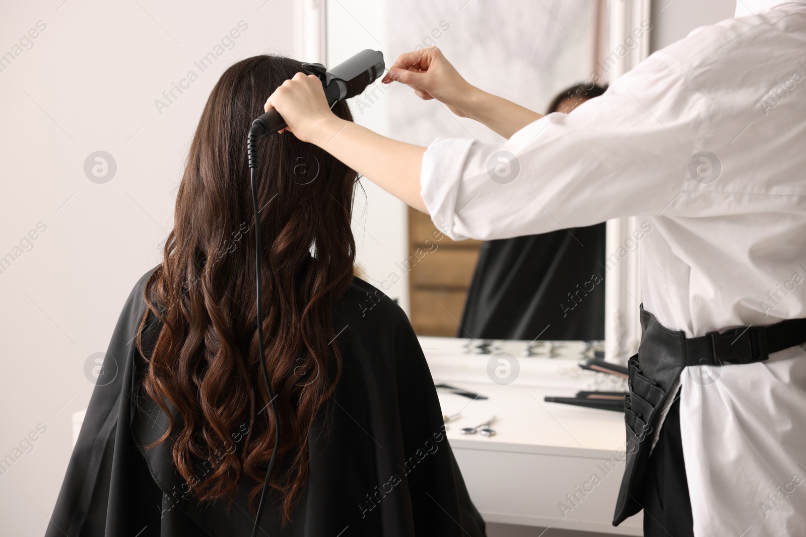 Photo of Hairdresser working with client using curling hair iron in salon, closeup