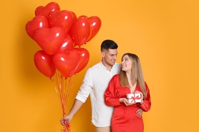 Photo of Lovely couple with heart shaped balloons and gift on yellow background. Valentine's day celebration