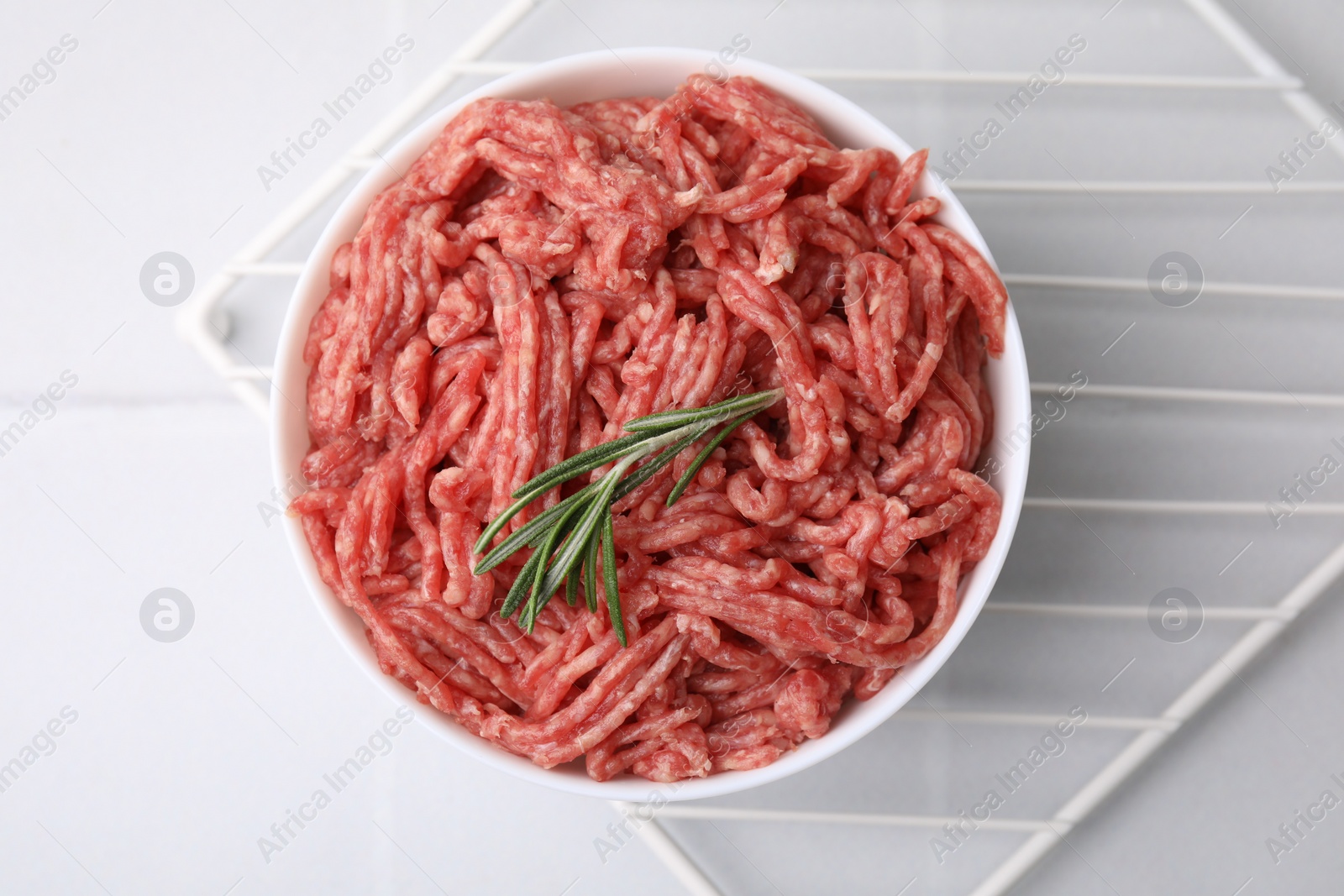 Photo of Fresh raw ground meat and rosemary in bowl on white tiled table, top view