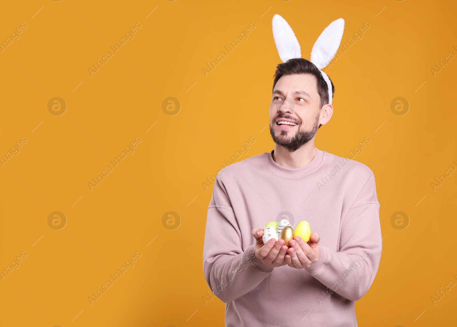 Photo of Happy man in bunny ears headband holding painted Easter eggs on orange background. Space for text