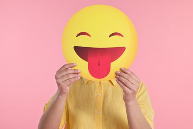 Photo of Woman covering face with emoticon sticking out tongue on pink background