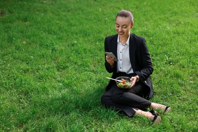 Photo of Lunch time. Happy businesswoman with container of salad using smartphone on green grass outdoors, space for text
