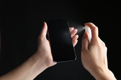 Photo of Woman spraying antiseptic onto smartphone against black background, closeup