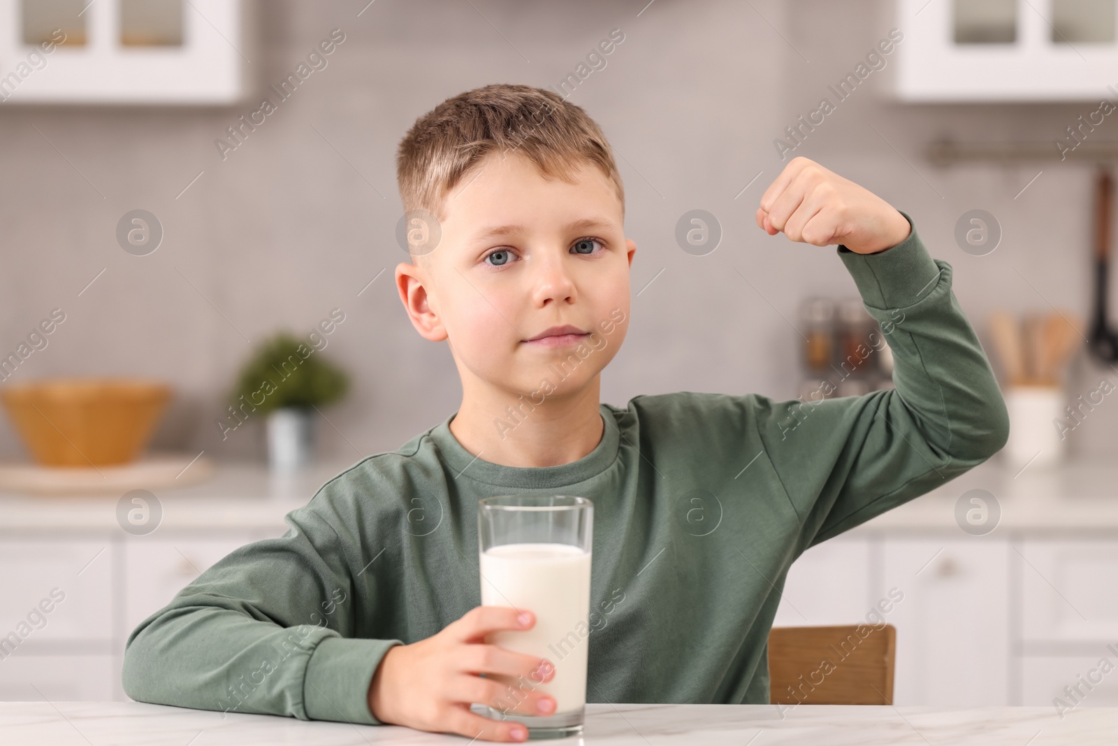 Photo of Cute boy with glass of fresh milk showing his strength at white table in kitchen
