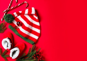 Photo of Cute striped elf hat, baby booties and Christmas decorations on red background, flat lay. Space for text