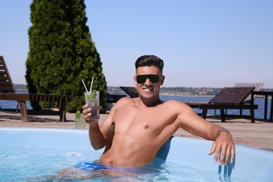 Happy man with cocktail in outdoor swimming pool on sunny summer day