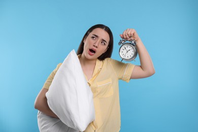 Photo of Tired young woman with pillow and alarm clock yawning on light blue background. Insomnia problem