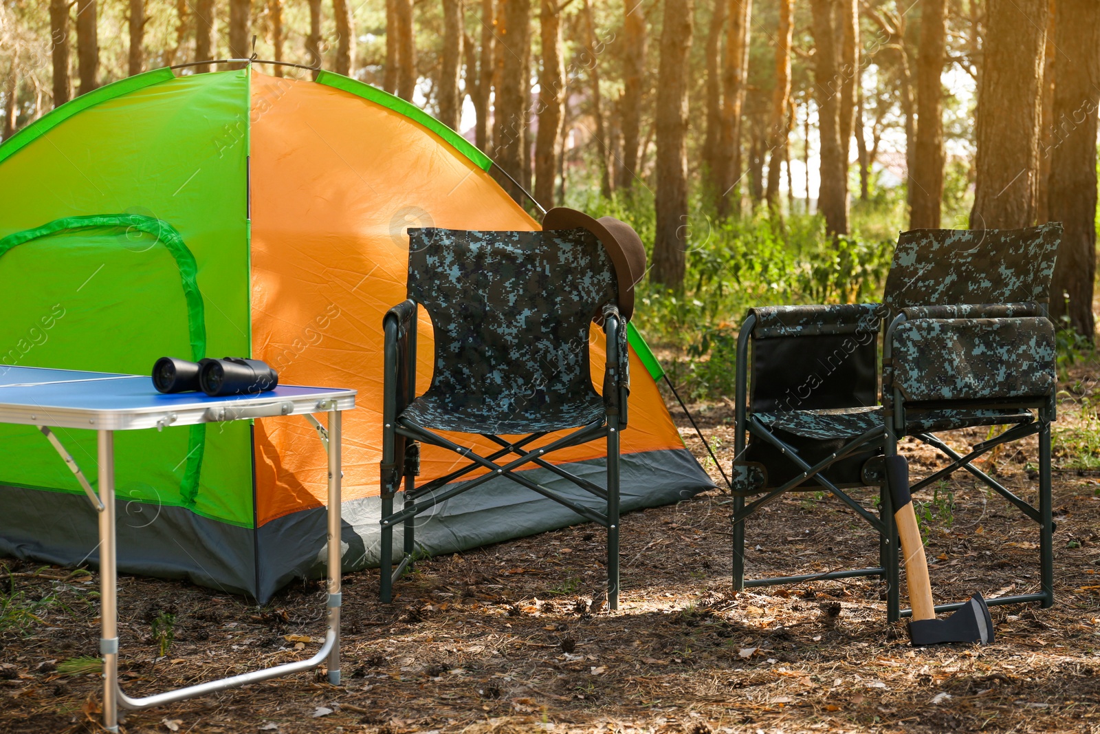 Photo of Camouflage chairs with hat, axe and table near camping tent in forest on sunny day