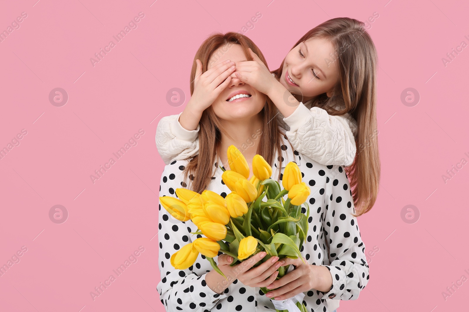 Photo of Daughter covering mother's eyes with her palms on pink background, space for text. Woman holding bouquet of yellow tulips