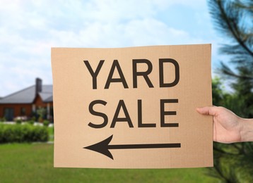 Image of Woman holding sign with text YARD SALE and blurred view of modern house on sunny day