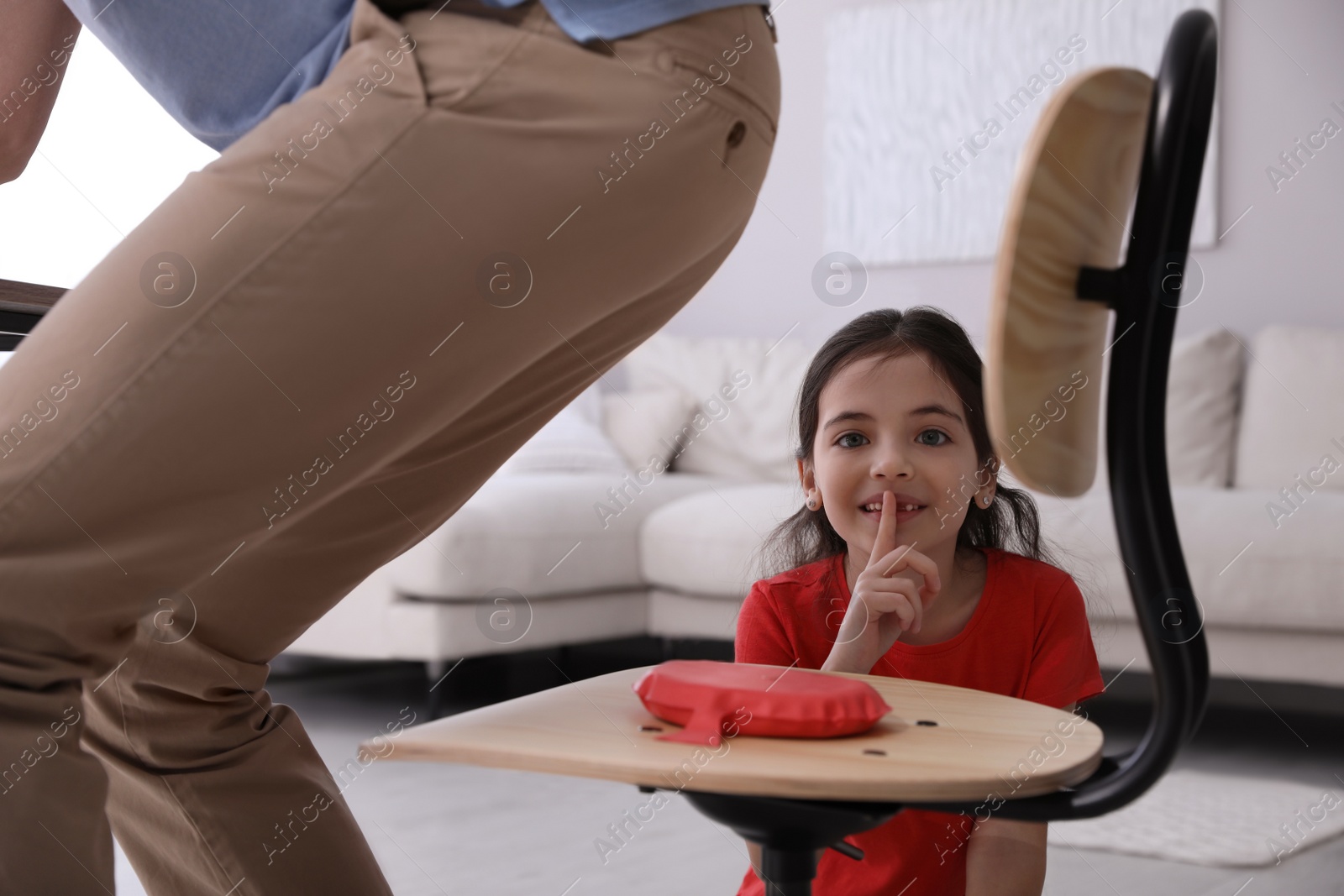 Photo of Cute little girl putting whoopee cushion on father's chair while he sitting down at home, closeup