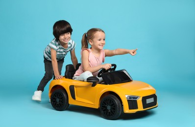 Photo of Cute boy pushing children's electric toy car with little girl on light blue background