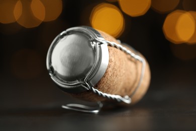 Sparkling wine cork with muselet cap on black table against blurred festive lights, closeup