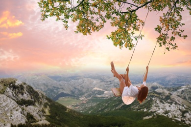 Image of Dream world. Young woman swinging over mountains under sunset sky 