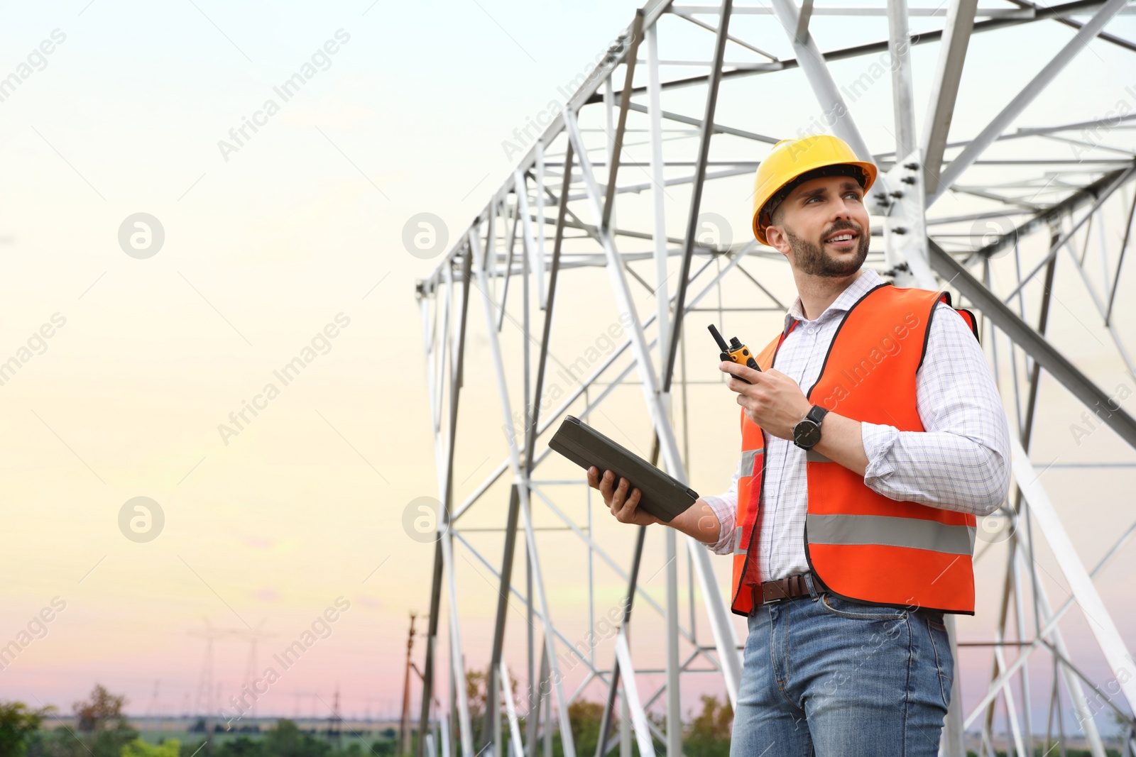 Photo of Professional engineer with tablet and walkie talkie near high voltage tower construction outdoors. Installation of electrical substation