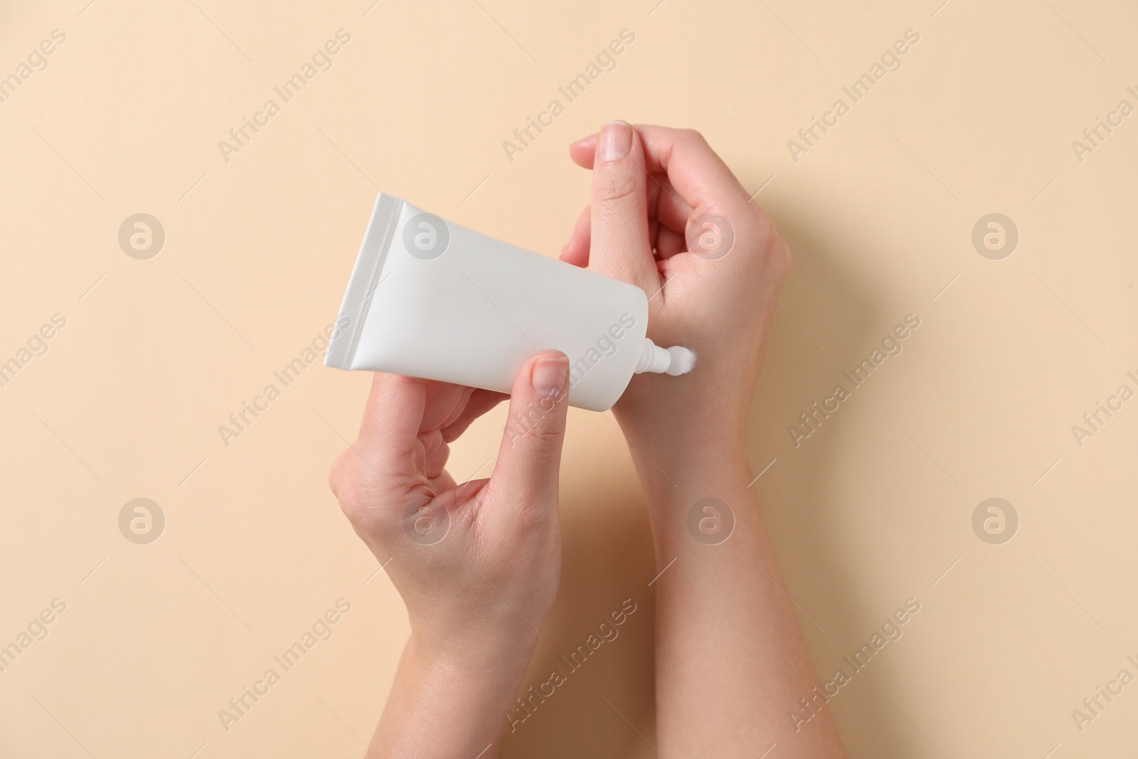 Photo of Woman applying cosmetic cream from tube onto her hand on beige background, top view