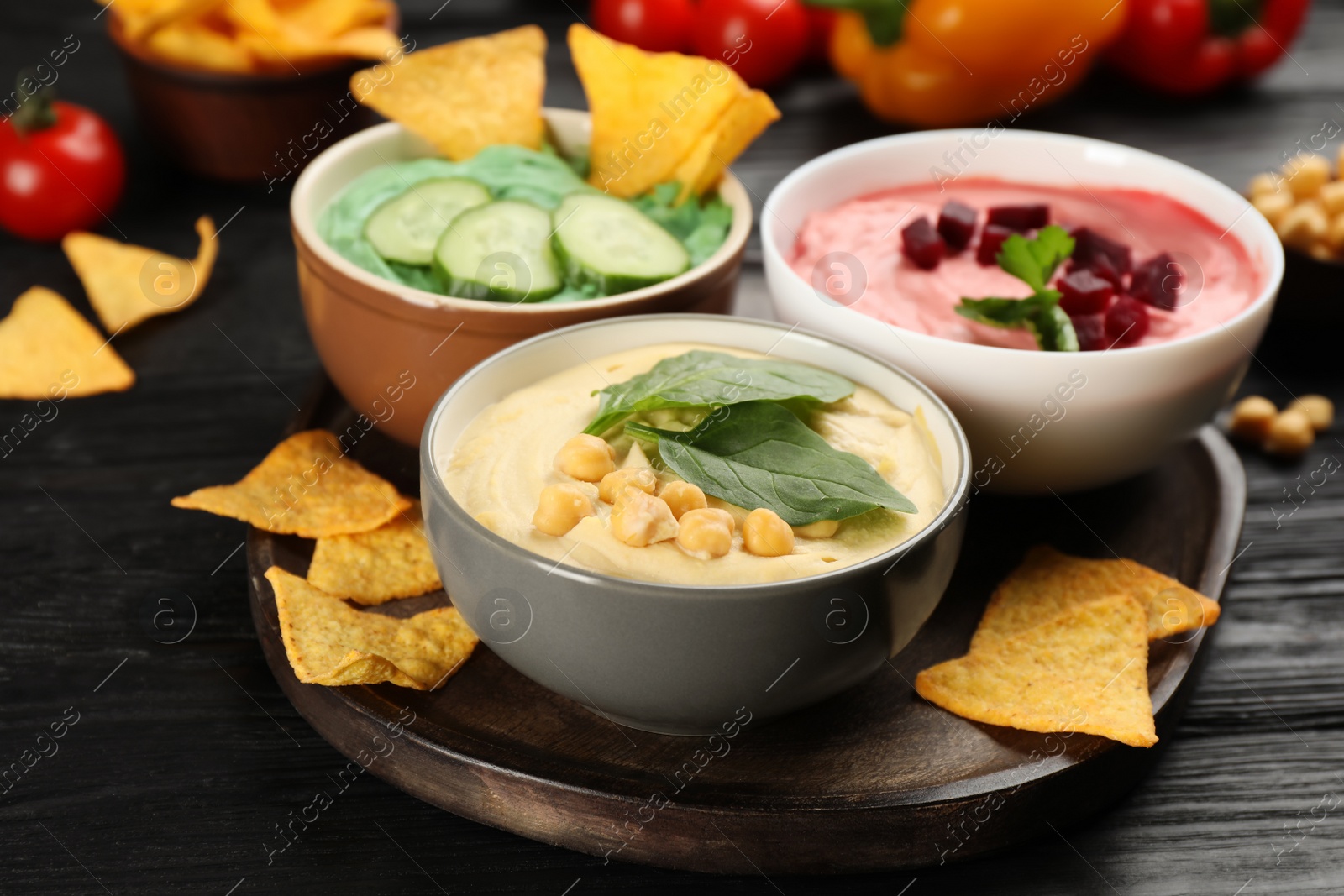 Photo of Different kinds of tasty hummus, nachos and ingredients on black wooden table