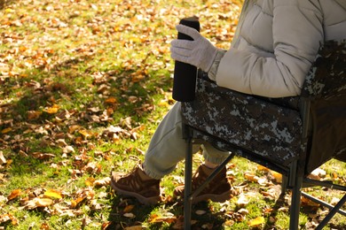 Photo of Woman with thermos sitting in camping chair outdoors on autumn sunny day, closeup