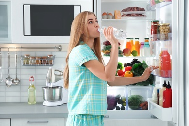 Woman drinking water out of bottle near refrigerator in kitchen