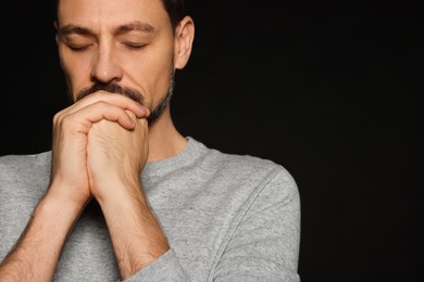 Photo of Man with clasped hands praying on black background, closeup. Space for text