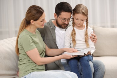Girl and her godparents reading Bible together on sofa at home