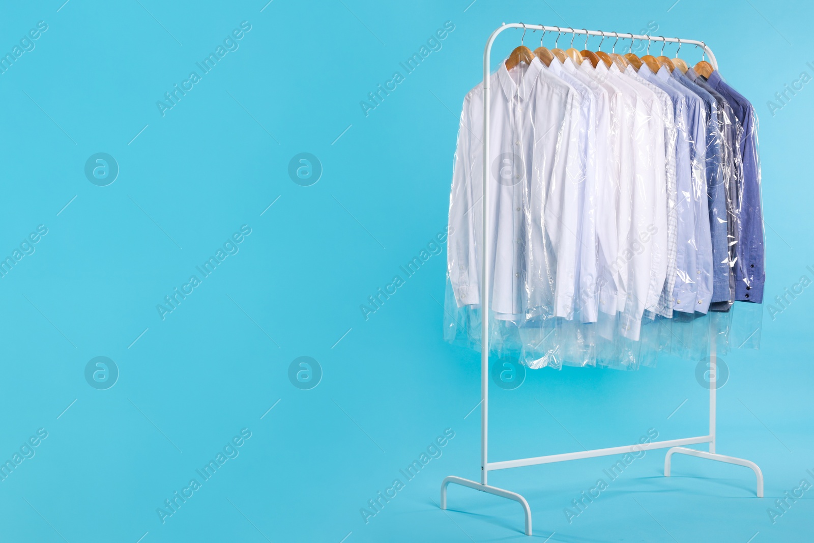 Photo of Dry-cleaning service. Many different clothes in plastic bags hanging on rack against light blue background, space for text