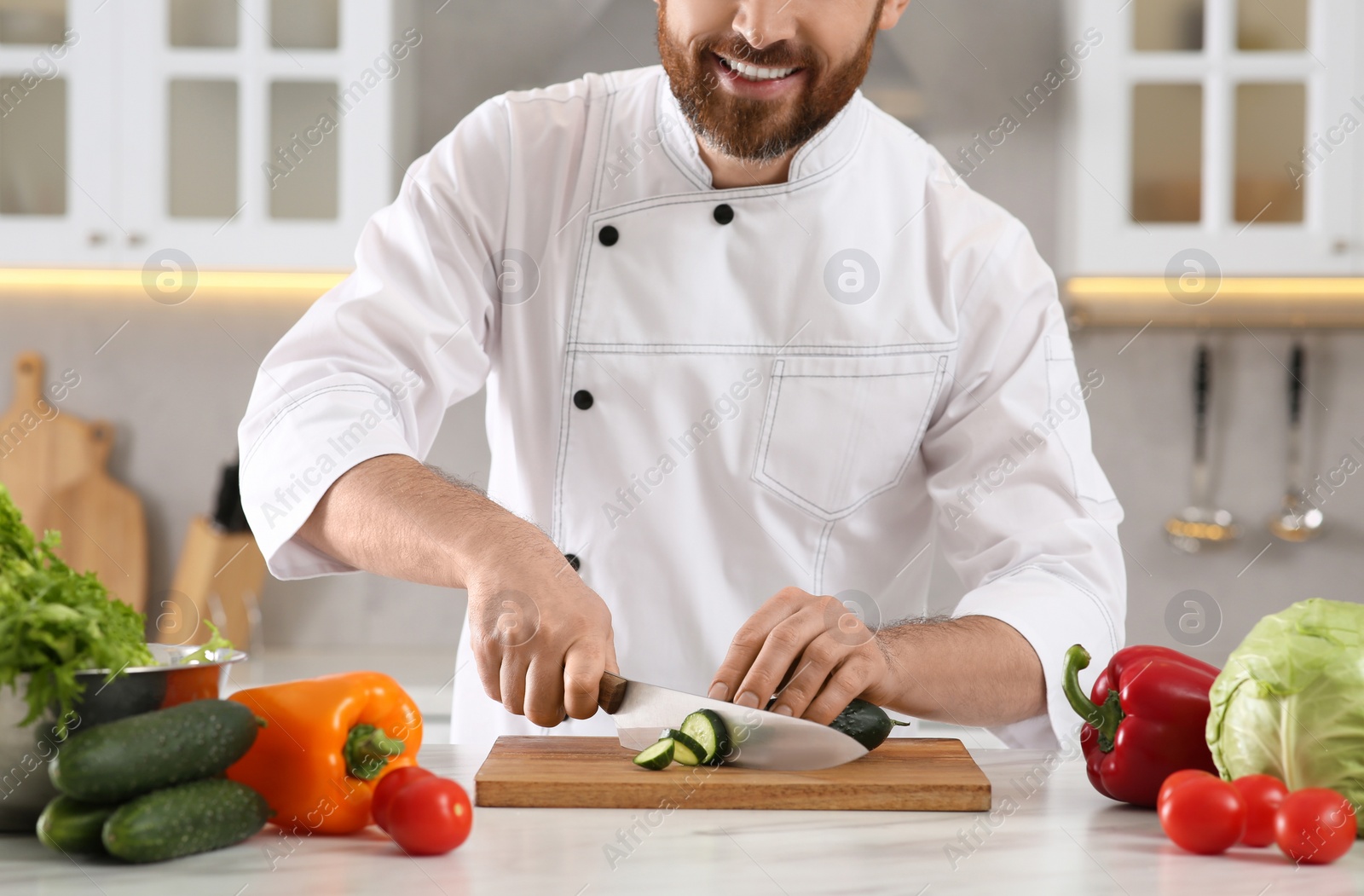 Photo of Happy chef cutting cucumber at marble table in kitchen, closeup