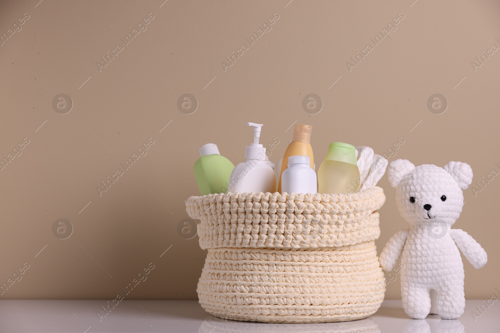 Photo of Knitted basket with baby cosmetic products and toy bear on white table against beige background. Space for text
