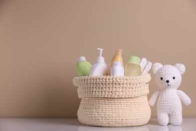 Photo of Knitted basket with baby cosmetic products and toy bear on white table against beige background. Space for text