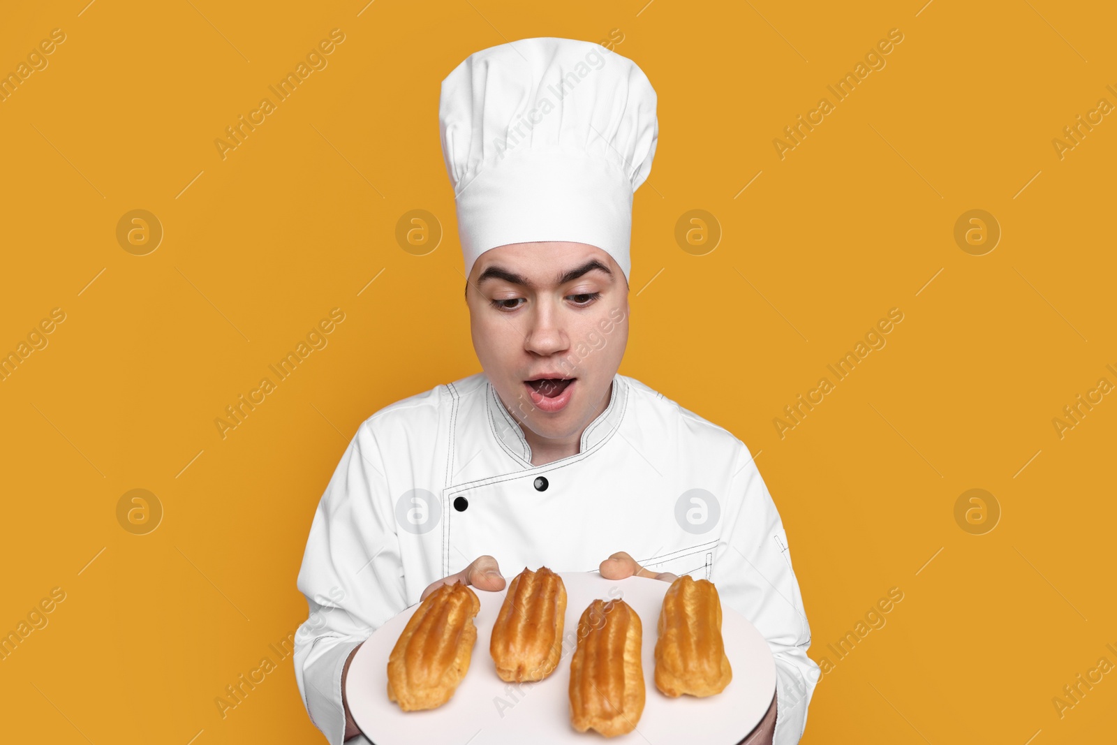 Photo of Portrait of emotional confectioner in uniform holding plate with eclairs on orange background