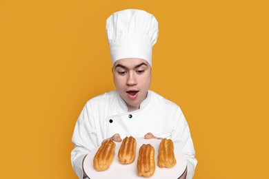 Portrait of emotional confectioner in uniform holding plate with eclairs on orange background