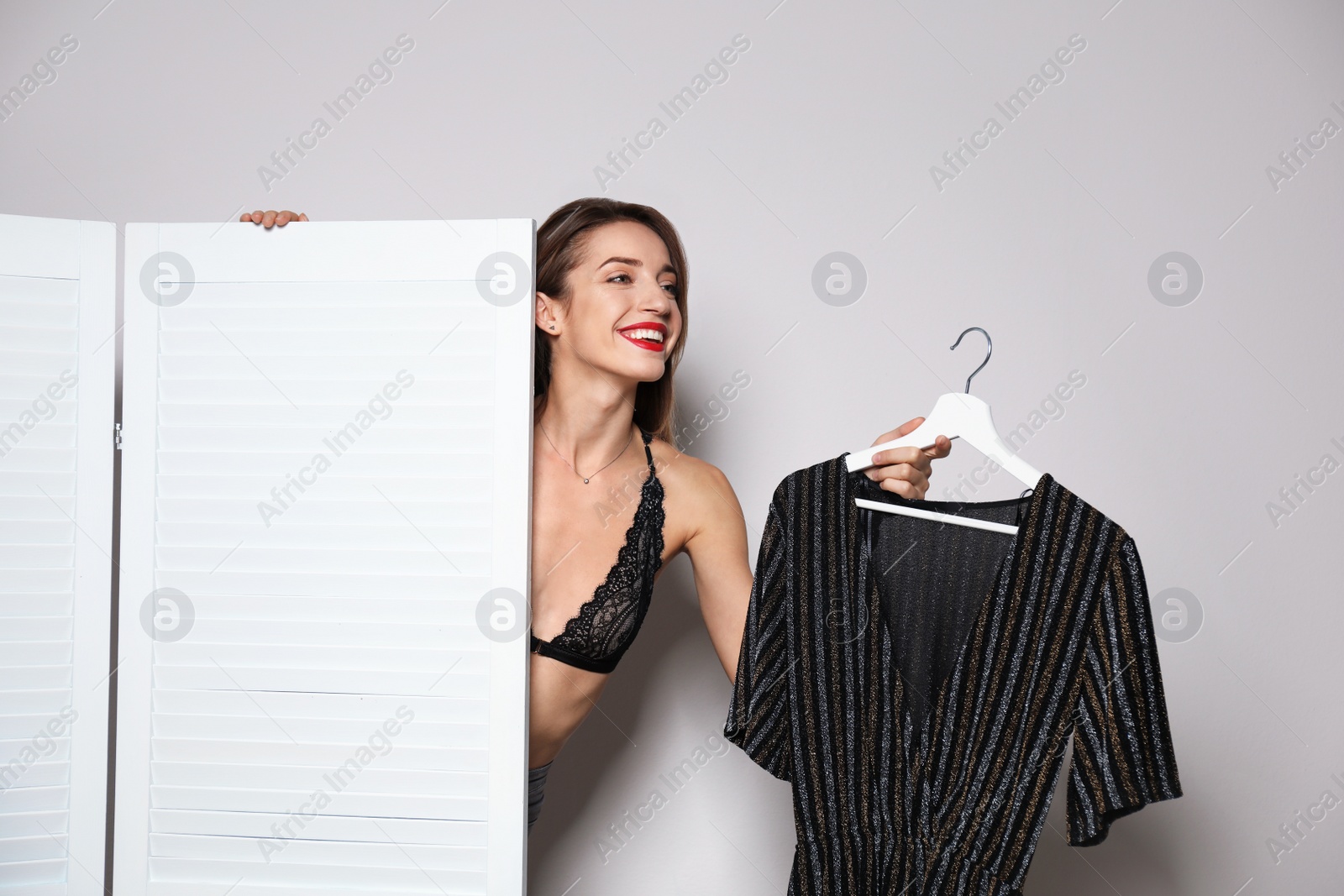 Photo of Young woman with clothes on hanger behind folding screen against light background. Dressing room