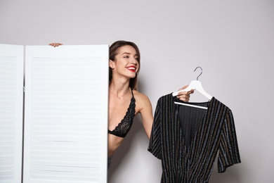 Young woman with clothes on hanger behind folding screen against light background. Dressing room