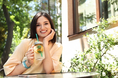 Photo of Young woman with mason jar of tasty natural lemonade at table, outdoors. Detox drink