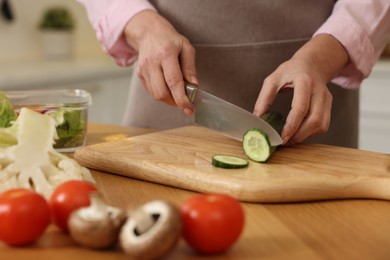 Woman cutting cucumber at wooden table in kitchen, closeup