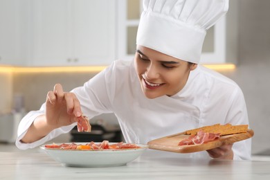 Photo of Professional chef adding prosciutto into delicious dish at white marble table in kitchen